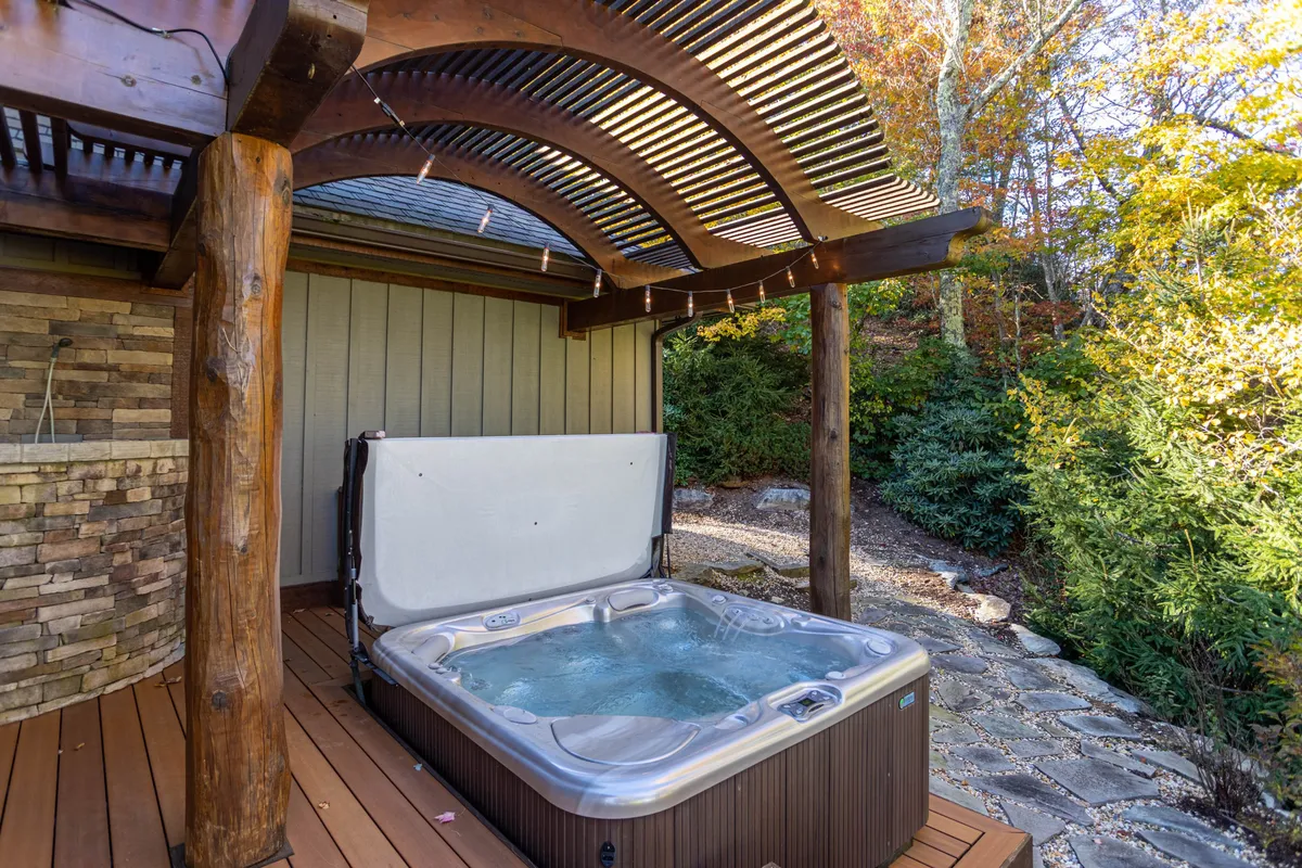 This image features a tranquil outdoor area with a hot tub under a wooden pergola and a stone wall, set against a natural backdrop. The setting invites relaxation and stargazing, delivering on the promise of leisure and luxury after a day of exploring local attractions.