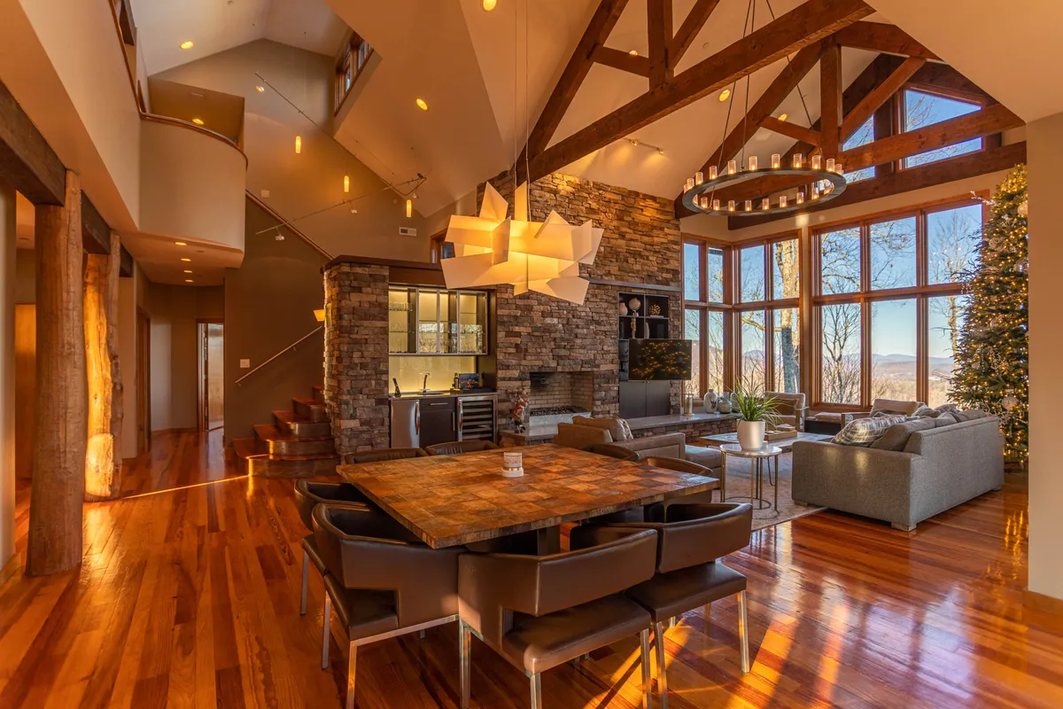 An elegantly designed open-concept living and dining area, featuring expansive windows that frame lush outdoor vistas. The space includes a modern dining table, a striking stone fireplace, and stylish light fixtures. The layout is ideal for entertaining, with seamless transitions between relaxation and dining zones.