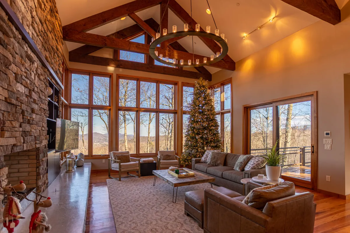A spacious main living room with a large, rustic stone fireplace as the central feature. Tall, floor-to-ceiling windows overlook a sweeping landscape, allowing natural light to flood the room. Plush, comfortable seating is arranged to encourage conversation or cozy relaxation by the fire. A chandelier adds a touch of elegance, and the mood is both warm and inviting, perfect for gatherings.