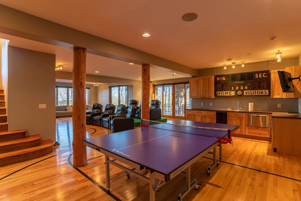 A lively recreation room featuring a ping pong table, comfortable seating, a kitchenette, and large windows letting in natural light. The space is set up for active play, relaxation, and socializing with friends or family.