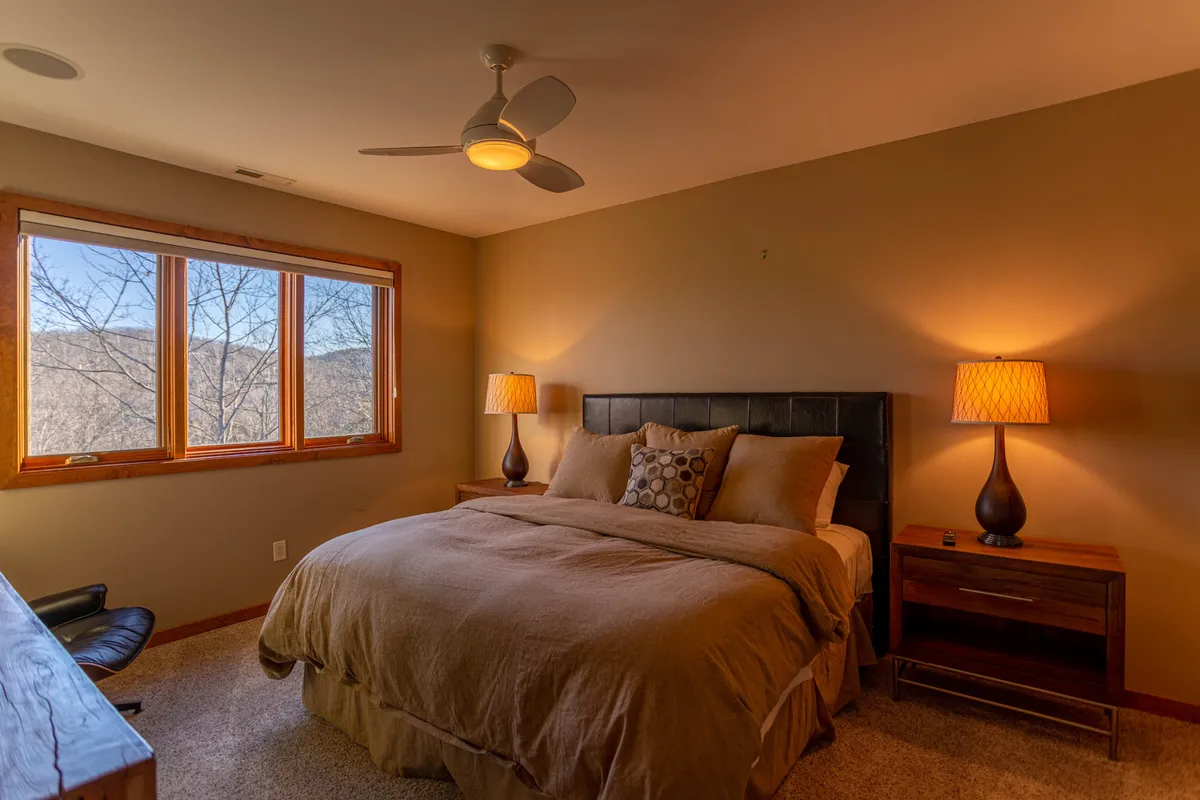 A tranquil bedroom with a plush bed adorned in decorative pillows, large windows offering a picturesque mountain view, and nightstands with soft lighting. The atmosphere is serene and restful.