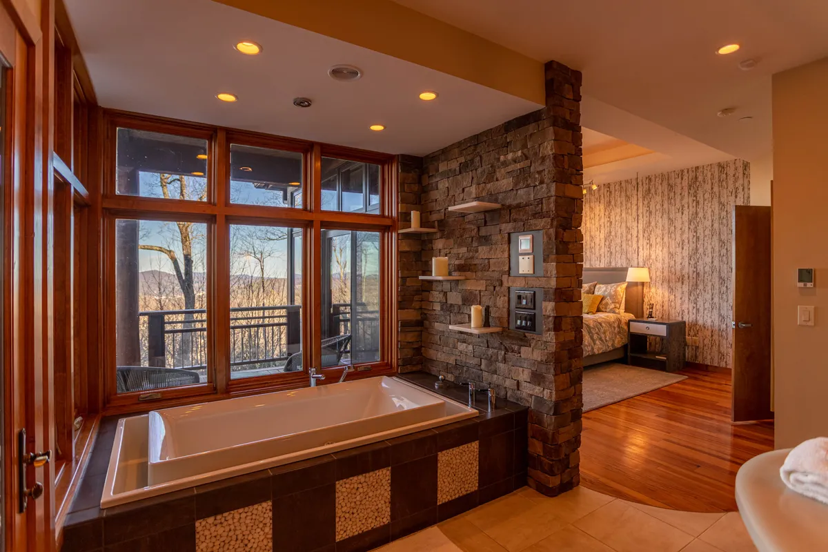 An inviting bathroom with a deep soaking bathtub placed beside large windows, a stone wall, and the entrance to a connecting bedroom. The space is bathed in natural light and overlooks scenic outdoor views.