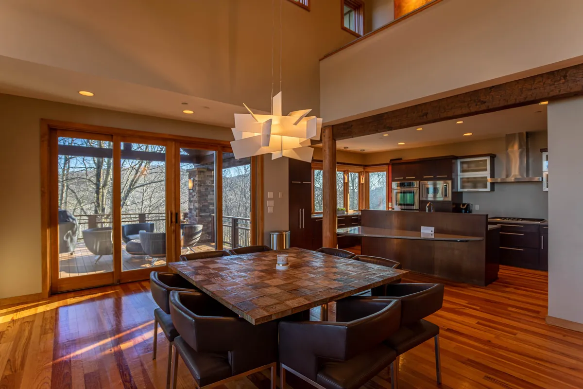 A stylish dining area adjacent to the kitchen, with a long dining table, modern light fixture, and large windows letting in abundant sunlight. The kitchen area is visible in the background, connecting the two spaces.