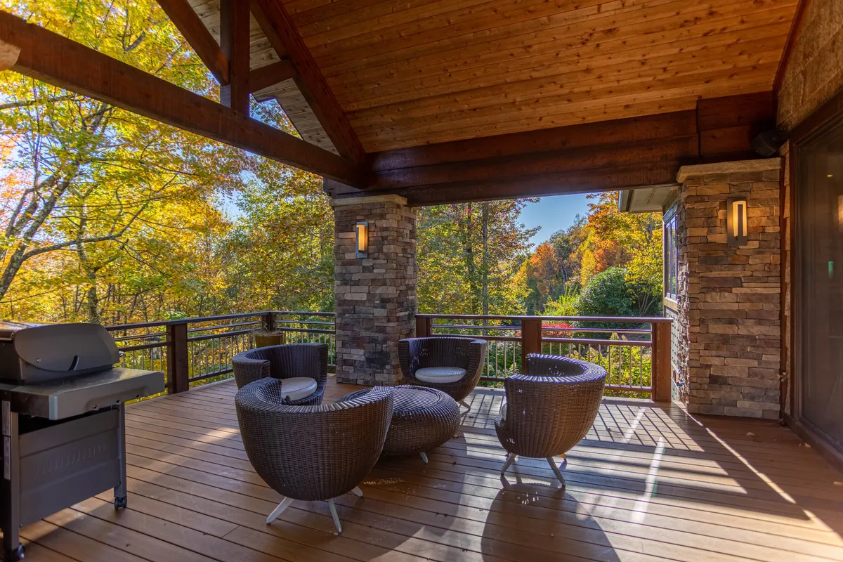 A covered outdoor deck with wooden ceiling beams, stone accents, and woven seating. The setup includes an outdoor grill and is surrounded by foliage, creating a serene, inviting spot for social gatherings. The space feels like a natural extension of the indoor living areas.