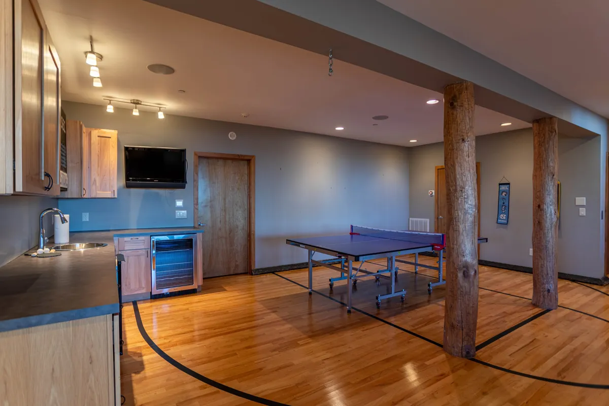 An energetic recreation space with a ping pong table, wooden pillars, and a convenient kitchenette area. The layout encourages friendly competitions, casual gatherings, and easy access to snacks or drinks.