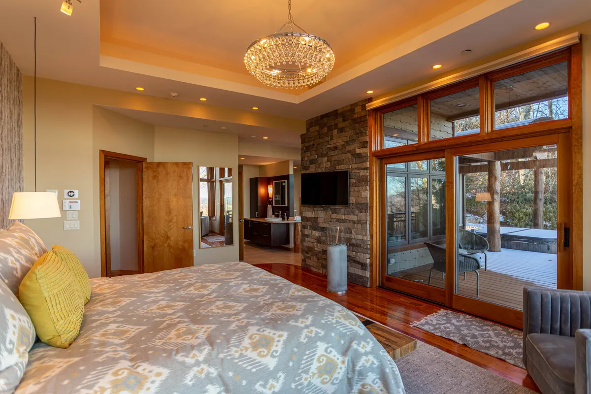 A luxurious bedroom featuring a large bed with decorative pillows, a striking chandelier overhead, expansive windows letting in natural light, and a stone accent wall. The room exudes both comfort and elegance, ideal for a master suite.