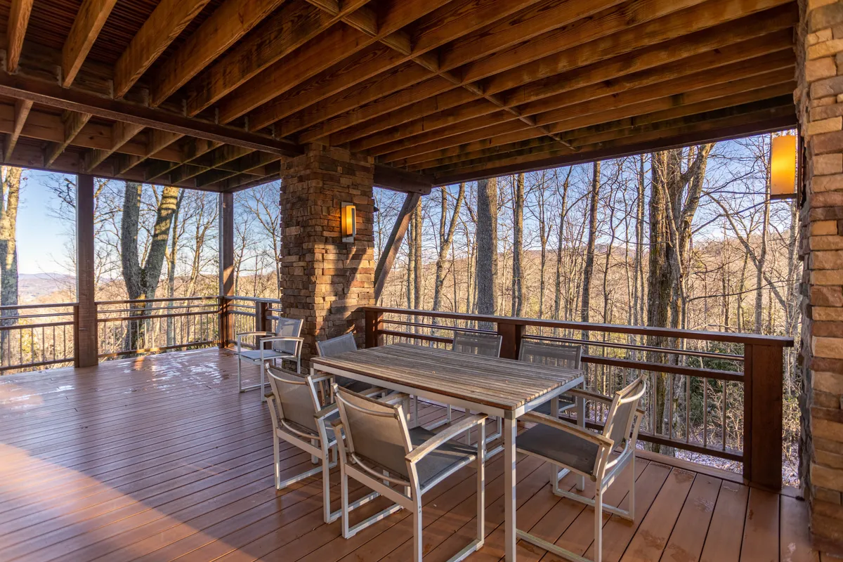 The image features a wooden deck with an outdoor dining table surrounded by trees, stone walls, and warm lighting fixtures. The space is designed for outdoor gatherings, nature watching, and relaxing, with a tranquil, welcoming ambiance.